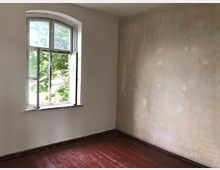 Ein leerer Raum mit abgeplatzter Putzwand auf der rechten Seite, einer geöffneten Rundbogenfenster mit Blick auf grüne Bäume und einem Boden aus dunklem Holz.