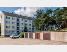 Mehrfamilienhaus mit vier Etagen und hellem Anstrich, flankiert von Bäumen auf der rechten Seite. Im Vordergrund stehen eine Reihe verschlossener Garagen sowie ein geparkter Pkw auf einem gepflasterten Hof.