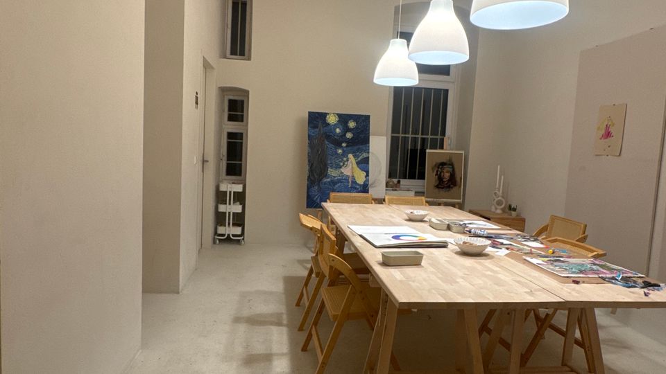 Langer, heller Atelierraum mit hohen weißen Wänden und Betonboden, in der Mitte ein großer Holztisch mit mehreren Holzstühlen und ausliegenden Mal- und Bastelmaterialien. Über dem Tisch hängen weiße Pendelleuchten, im Hintergrund stehen mehrere Gemälde an der Wand und bei einem Fenster.