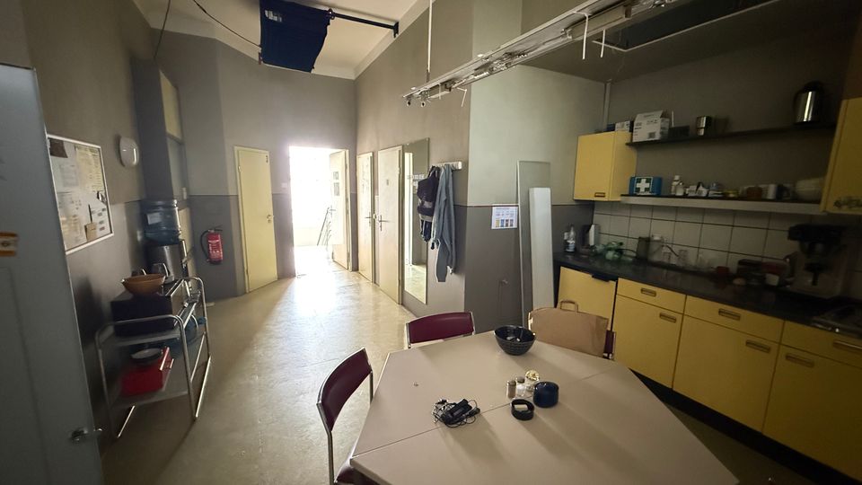 Schmaler Pausenraum mit Tisch und Stühlen im Vordergrund sowie einer Küchenzeile mit gelben Schränken, Spüle und offenen Regalen an der rechten Wand. Am Ende des Raums führen mehrere Türen in angrenzende Bereiche; an der linken Seite stehen ein Wasserspender und ein Servierwagen.