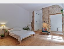 Schlafzimmer mit hellem Holzboden, einem einfachen Holzbett mit weißer Bettwäsche und mehreren Zimmerpflanzen. Eine unverputzte Backsteinwand mit offener Durchgangsöffnung und ein blauer Sessel mit bunter Decke setzen Akzente, während eine Stehlampe für Beleuchtung sorgt.