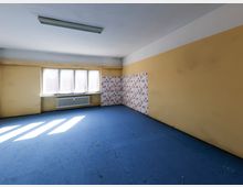 Leerer Innenraum mit blauem Boden, beigefarbenen Wänden und großer Fensterfront mit Heizkörper darunter. An einer Ecke befindet sich ein Abschnitt mit rot gemusterter Tapete, an der Decke hängt eine lange Leuchtstofflampe.