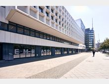 Modernes Gebäude im Baustil der Nachkriegsmoderne mit einer Fassade aus weißen rechteckigen Elementen und großen Glasflächen; im Vordergrund ein breiter, gepflasterter Gehweg mit vereinzelten Personen. Im Hintergrund sind weitere moderne Gebäude und ein Turm erkennbar.