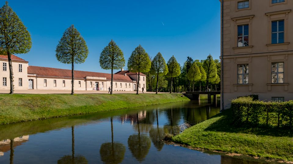 Canal bordered by grassy banks and a row of evenly spaced, trimmed trees, with long beige buildings topped by red tile roofs along the water. A larger beige building with tall windows frames the right side, and a small bridge crosses the canal in the distance under a clear blue sky.