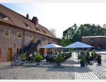Ein Backsteingebäude mit roten Ziegeln und einem Satteldach, dessen Fassade von mehreren rechteckigen Holzfenstern und einer Außentreppe geprägt ist. Davor befindet sich ein gepflasterter Hof mit Sitzplätzen, Sonnenschirmen und Pflanzen in Töpfen, umgeben von weiterer Begrünung und Nebenbauten.