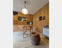 Holzvertäfelter Raum mit einem Esstisch aus Holz, einem geflochtenen Korbhocker und einer Hängelampe mit dekorativem Glasschirm. Im Hintergrund befinden sich Holzschränke mit Glaseinsätzen sowie eine Heizung an der rechten Wand.