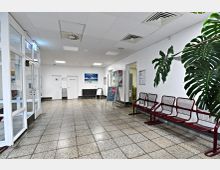 Ein heller, schlicht eingerichteter Warteraum mit roten Metallstühlen an der rechten Wand, großen Bodenfliesen und weißen Wänden. Links befindet sich eine Glastür, in der Mitte Türen zu Toiletten und „Kein Zutritt“, sowie ein kleiner Heizkörper unter einem Bild mit Strandmotiv.