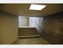 Unterführung mit gefliesten Wänden und Treppen, die zu einem metallenen Rolltor hinaufführen. An der Decke befindet sich eine quadratische Beleuchtung, und ein Geländer verläuft entlang der rechten Wand.