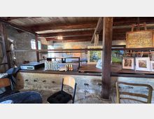 Innenraum eines rustikalen Steingebäudes mit sichtbaren Holzbalken und Holzdecke, in dem auf einem langen Tresen gerahmte Gemälde und Drucke ausgestellt sind. Im Vordergrund stehen Stühle und ein Tisch, daneben befinden sich Flaschen und Gläser auf dem Tresen; kleine Fenster und Holztüren prägen den Raum.