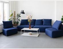 Helles Wohnzimmer mit großen Fenstern, weißen Wänden und grauem Boden, eingerichtet mit einem modularen, dunkelblauen Sofa und passendem Sessel. Davor steht ein niedriger weißer Couchtisch mit Blumen in einer Vase; daneben eine schwarze Stehlampe und mehrere Zimmerpflanzen.