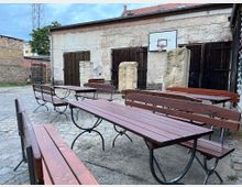 Ein gepflasterter Innenhof mit mehreren Holzbänken und Tischen. Im Hintergrund befinden sich eine verwitterte Hauswand mit dunklen Holztüren, ein Basketballkorb und zwei hohe Steinblöcke.