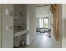 Schlichter Innenraum mit grauem Betonboden und weißen Wänden. Im Vordergrund befindet sich ein schmaler Waschbereich, im Hintergrund ein Wohnbereich mit grauem Sofa, Glastisch und großem Fenster mit weißen Vorhängen.