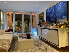 Ein gemütliches Wohnzimmer mit modernen Möbeln, einem weißen Sofa, einem flauschigen Teppich und einem niedrigen Couchtisch. Im Hintergrund ist eine Terrassentür mit dekorativer Beleuchtung, und auf der Kommode stehen weihnachtliche Dekorationen neben einem großen Fernseher an der Wand.