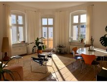 Heller Wohnraum mit großen, bogenförmigen Fenstern, die von weißen Vorhängen umgeben sind. Der Raum enthält eine Sitzecke mit einem schwarzen Designersessel, einem Beistelltisch, Zimmerpflanzen, einer runden Esstischgruppe mit orangefarbenen Stühlen und einem Holzfußboden.