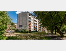 Wohnblock mit fünf Etagen, bunten Balkonen und schlichtem Fassadendesign in einer grünen Umgebung mit Bäumen, Wiese und einem kleinen Spielplatz im Vordergrund. Im Hintergrund sind weitere Wohngebäude sichtbar.