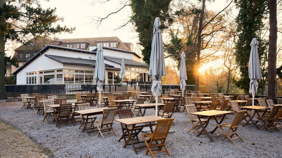 Outdoor café patio with many wooden tables and folding chairs on a gravel surface, dotted with tall closed umbrellas. A low white building with large windows sits behind the seating area, surrounded by tall trees with sunlight filtering through.