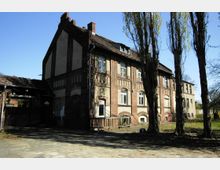 Zweistöckiges historisches Gebäude aus Backstein mit hohen Fenstern und dekorativen Elementen an der Fassade; daneben ein kleiner überdachter Eingangsbereich und Bäume im Vordergrund. Im Hintergrund ist ein weiteres schlichtes Gebäude und freies Gelände erkennbar.