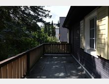 Holzterrasse eines Gebäudes mit dunkler Holzfassade, gelben Fensterläden und einem Geländer aus Holz. Im Hintergrund sind dichte Bäume, eine Nachbarfassade mit Ziegeldach und ein blauer Himmel sichtbar.