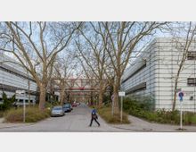 Straßenszene mit modernen Bürogebäuden aus Metall und Glas auf beiden Seiten, dazwischen Gehwege mit großen, kahlen Bäumen. Im Hintergrund verbindet eine gläserne, rot umrahmte Fußgängerbrücke die Gebäude.