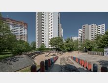 Wohngebiet mit mehreren Hochhäusern, umgeben von Bäumen und Grünflächen. Im Vordergrund befindet sich ein Spielplatz mit Sandboden, Spielgeräten aus Holz sowie einem gepflasterten Weg mit rot-schwarzen Pollern.
