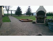 Gepflasterter Hofbereich mit einer gemauerten Gartenfeuerstelle, darunter gestapeltes Holz. Im Hintergrund eine Rasenfläche mit zwei Nadelbäumen, einem Sandkasten und einem Zaun, der das Gelände abgrenzt.