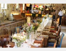 Langer, rustikaler Holztisch in einem gemütlichen Innenraum, gedeckt mit Tellern, Gläsern und Blumenarrangements; im Hintergrund eine beleuchtete Bar und unscharfe Personen.