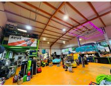 Große Autowerkstatt mit hoher Hallendecke und sichtbaren Trägern, orangem Boden und zahlreichen Werkzeugen sowie Regalen. Mehrere Oldtimer stehen oder hängen auf Hebebühnen; an der Decke ist eine rechteckige, pink beleuchtete Konstruktion angebracht.