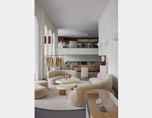 Modernes Innendesign eines Showrooms mit hellbeigen, geschwungenen Sesseln und einem ovalen Couchtisch auf einem Teppich. Im Hintergrund befinden sich Kleidung auf Ständern, weitere Sitzmöbel, ein Regal und eine Galerie mit Glasgeländer.