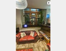 Gemütliche Sitzecke in einem Innenraum mit rotem, geknöpftem Leder-Chesterfield-Sofa vor einem großen Holzrahmenfenster. Daneben stehen ein Klavier mit ausliegenden Zeitschriften, ein kleiner Hocker, Zimmerpflanzen sowie Dekoration auf der Fensterbank.