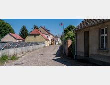 Kopfsteingepflasterte Dorfstraße mit einseitig stehenden Häusern im traditionellen Stil, darunter gelbe und rote Dächer. Links verläuft ein Holzzaun mit Gewächshäusern dahinter, rechts ein altes Gebäude und eine rote Straßenlaterne.