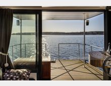 Blick aus dem Inneren eines Hausbootes durch eine Glastür auf eine Holzterrasse und einen See mit glitzernder Wasseroberfläche. Im Hintergrund sind bewaldete Ufer zu sehen, während die Terrasse mit einer Reling umgeben ist.