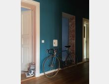 Ein Innenraum mit einer Wand in dunklem Blau, an der ein blaues Rennrad lehnt. Links befindet sich eine geöffnete weiße Tür mit Zierleisten, daneben ein Stapel Bücher und CDs, rechts ist teilweise unverputztes Mauerwerk sichtbar. Der Boden besteht aus Holz.