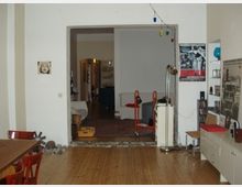 Wohnzimmer mit Holzboden und Blick in einen angrenzenden Raum. Im Vordergrund ein Esstisch mit roten Stühlen, an der rechten Wand ein Sideboard mit Dekorationen, und im Hintergrund Wohnaccessoires sowie teilweise sichtbare Möbel.