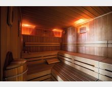 Innenansicht einer Sauna aus Holz mit mehreren Sitz- und Liegebänken, dezent beleuchtet durch warme Lichtquellen an den Wänden. Im Vordergrund ist ein hölzerner Eimer abgebildet, und die Wände und Decke bestehen vollständig aus Holzvertäfelung.