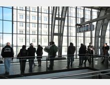 Innenbereich eines Bahnhofs mit großen Glasfassaden und Metallträgern, durch die ein benachbartes Gebäude sichtbar ist. Mehrere Menschen stehen oder gehen auf dem Bahnsteig im Wartebereich, einige mit Taschen oder einer Zeitung in der Hand.