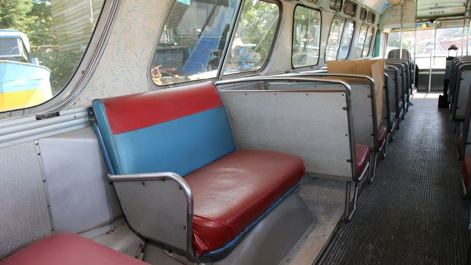 Innenraum eines alten Linienbusses mit rot-blauen Sitzen und Metallrahmen. Große Fenster an den Seiten lassen Licht herein, und der Boden besteht aus dunklem geriffelten Material.