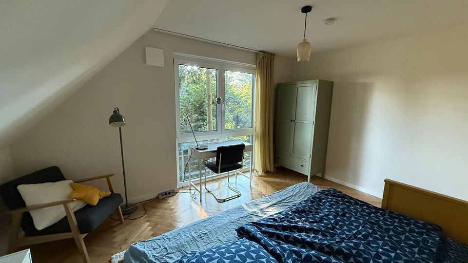 Ein helles Schlafzimmer mit Dachschräge, einem großen Fenster mit Blick auf Bäume, einem Bett mit blauer Bettdecke, einem Schreibtisch mit Stuhl, einem Sessel und einem grünen Kleiderschrank. Der Boden ist aus Holzparkett, und eine Pendelleuchte hängt an der Decke.