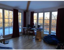 Ein Wohnzimmer mit großen bodentiefen Fenstern, durch die ein Balkon mit Geländer und schneebedeckte Bäume sichtbar sind. Der Raum hat Holzfußboden, rote Vorhänge, einen Schreibtisch mit Stuhl, eine kleine Kommode und lose Gegenstände wie einen Sitzsack.