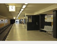U-Bahn-Station mit einem überdachten Bahnsteig, Sitzbänken, einer Wanduhr und Wegweisern; im Hintergrund ein Ausgang mit Tageslicht und Personen, die sich auf dem Bahnsteig bewegen. Ein Schild kennzeichnet die Station als „Ullsteinstraße“.