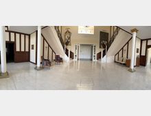 Weitläufiger Eingangsbereich mit Marmorboden, zwei geschwungenen Treppen mit schmiedeeisernen Geländern und weißen Säulen. Der Raum ist mit Wandvertäfelungen, Zierleisten, Spiegeln und Sitzmöbeln ausgestattet; im Hintergrund liegt eine Doppeltür unter einem hohen Fenster.