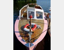 Ein kleines Holzboot mit rosa Rand liegt an einem Steg auf einem ruhigen Gewässer. Das Deck ist mit einem Rettungsring, Seilen und einer Lampe ausgestattet, und im Hintergrund sind teilweise geöffnete Fenster der Kabine sowie ein stehender Mann sichtbar.