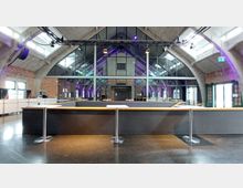 Innenraum eines modernen Veranstaltungsortes mit hohen Decken aus Beton, Backsteinwänden und einer großen Glasfront, die mehrere schwarze Stahlrahmen enthält. Im Vordergrund befindet sich ein Tresen mit Holzkante, während farbige Lichter die Rückwand und die Deckenbalken leicht beleuchten.