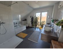 Modernes Badezimmer mit bodengleicher Dusche, weißer Badewanne und Doppelwaschbecken auf einem Holzunterschrank. Große Fenster mit Blick ins Freie lassen viel Tageslicht herein, ergänzt durch Zimmerpflanzen und dunkle Bodenfliesen.