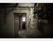 Dunkler Kellerraum mit bröckelnden Wänden, sichtbaren Rohren und Ventilen an der rechten Wand. Im Hintergrund führt eine Türöffnung zu einem weiteren Raum mit einem kleinen, vergitterten Fenster, das schwaches Licht hineinlässt.
