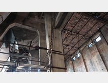 Innenansicht eines verlassenen Industriegebäudes mit hohen Backsteinwänden, einer teilweise zerstörten Decke aus Stahlträgern und Ziegeln sowie Metallleitern und Plattformen. Die spärlichen Fenster lassen Licht herein, und das Bauwerk zeigt deutliche Spuren von Verfall.