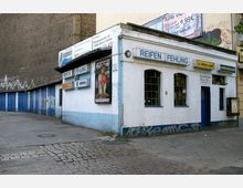 Kleine Werkstatt mit einem weißen Gebäude, blauen Fenstern und Werbeschildern für Reifenmarken wie Michelin und Dunlop; links im Bild sind eine Reihe blauer Garagentore zu sehen, während der Boden aus Beton und Kopfsteinpflaster besteht.