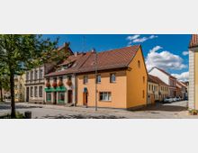 Straßenszene mit farbigen Wohn- und Geschäftshäusern, überwiegend mit roten Ziegeldächern. Im Vordergrund ein Gehweg mit einem Baum und Laternenpfahl, dahinter sind auf der Straße geparkte Autos zu sehen, bei klarem Himmel.