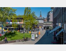 Blick auf den Bahnhof Berlin-Schöneberg mit einer erhöhten Bahntrasse, umgeben von Gebäuden und einem kleinen Platz mit Bäumen, Sitzgelegenheiten und Fahrradständern; im Hintergrund ist die Spitze einer Kirche sichtbar. Passanten und ein Bus prägen das belebte Stadtbild.