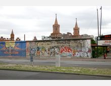 Ein Abschnitt der Berliner Mauer, bedeckt mit farbenfrohen Graffiti und Kunstwerken, im Vordergrund einer Straße. Im Hintergrund sind die Türme der Oberbaumbrücke zu sehen, eine markante rote Backsteinarchitektur.