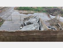 Ein verlassenes, teils eingestürztes Gebäude mit freiliegenden Mauern und Schutthaufen, bedeckt von grauen Planen. Der Boden ist stellenweise mit Wasser bedeckt, und das Gelände wirkt ungenutzt, umgeben von Vegetation und Trümmern.